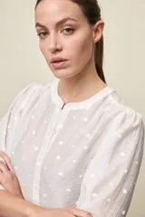Camisa negra de textura liviana con detalle de lunares en relieve, cuello mao y cierre frontal con botones. Presenta mangas 3/4 con puños elastizados y frunces en los hombros.