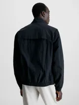 Campera negra de nailon arrugado reciclado, con cuello alto, puños y bajo elásticos, cierre con cremallera, bolsillos con cremallera en la cintura, abertura de ventilación en la espalda, forro de poliéster reciclado e insignia con el logo de goma de Calvin Klein en la manga.