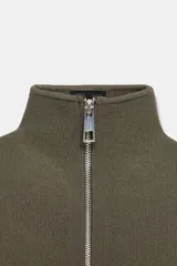 Chaqueta corta de tejido grueso color verde oliva, con cuello alto y cierre frontal completo de cremallera plateada. Presenta un corte amplio y mangas largas caídas.