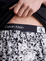 Conjunto de pijama Calvin Klein 1996, compuesto por una remera negra de algodón con cuello redondo, manga corta y logo estampado en el pecho, y un short de algodón tejido con estampado floral en blanco y negro, bolsillos laterales y cintura elástica con logo distintivo de la marca. Incluye bolsa de CK a juego.