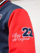 Campera bomber azul marino estilo retro, con cierre frontal de botones a presión, cuello, puños y ruedo con tejido acanalado tricolor. Presenta el logo de Umbro y el escudo del Club Nacional de Football bordados en el pecho, junto con el año 1899 y parches decorativos en las mangas.