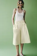 Bermudas de tiro medio color amarillo claro, confeccionadas en mezcla de algodón. Presentan cintura con trabillas, bolsillos laterales ocultos en costura, pliegues delanteros y cierre frontal con cremallera, botón y ganchos metálicos.