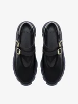 Zapato estilo bailarina con capellada de cuero negro charol y detalles en gamuza negra. Posee dos correas ajustables con hebillas metálicas plateadas sobre el empeine. La suela es gruesa y de goma, con diseño ondulado en color azul marino oscuro.