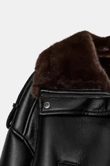 Cazadora bomber de efecto piel en color negro, con cuello subido, manga larga y bajo elástico. Presenta interior y puños con aplicación de pelo sintético en color marrón a contraste, cierre frontal con cremallera y botones a presión, y bolsillos laterales de vivo.