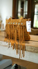 Cartera tipo bolso de cuero color tostado con diseño boho, decorada con flecos largos frontales y tachas metálicas circulares. Cuenta con doble asa de mano y cierre superior.