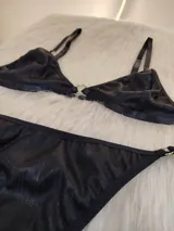 Conjunto de bikini color negro con textura acanalada. El corpiño es de estilo triangular con aros y detalle de argolla metálica en el centro. La bombacha es de corte colaless con tiras laterales ajustables y detalles de argollas metálicas.
