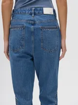 Pantalón de denim color marrón, de tiro alto y calce ochentoso con terminación estilo oxford. Cuenta con apliques metálicos decorativos alrededor de los bolsillos y a lo largo de las costuras exteriores.