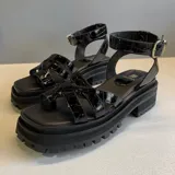 Sandalias de cuero sintético negro con efecto croco, suela flatform track dentada y tiras cruzadas en el empeine con sujetador para el dedo y tira ajustable al tobillo con hebilla metálica dorada.
