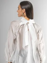 Camisa blanca de algodón italiano con cuello alto ajustable mediante lazo trasero, mangas abullonadas y un gran shabot frontal con volados en cascada.