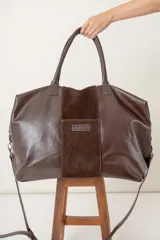 Bolso de viaje marrón de cuero vacuno con panel central de gamuza. Tiene doble asa de mano, correa al hombro regulable y desmontable, cierre superior y base con cuatro soportes metálicos.