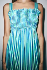 Vestido midi de escote recto con cuerpo de nido de abeja elástico, tirantes anchos con lazos y falda larga con volados en el borde inferior. Presenta un estampado de rayas verticales en tonos verde, azul y blanco.