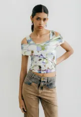 Top corto de jersey con estampado abstracto floral en tonos pastel (blanco, lila, verde, marrón). Presenta un diseño cruzado asimétrico en el busto que crea un escote tipo *off-shoulder* en un lado y manga corta en el otro.