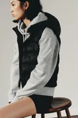 Chaleco acolchado de color negro con cuello subido, capucha ajustable y cierre frontal con cremallera termosellada. Presenta un diseño con paneles laterales en tejido liso y bolsillos laterales.