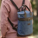 Mochila matera de diseño vertical, confeccionada en tela canvas marrón con detalles y refuerzos en cuero sintético oscuro. Cuenta con doble asa superior de mano, correas traseras regulables, cierre metálico frontal y bolsillo posterior.