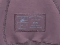 Buzo blanco de manga larga con diseño holgado, cuello redondo y costuras externas visibles en contraste. Cuenta con una etiqueta decorativa en el frente inferior.