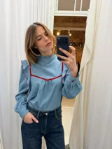 Blusa de jean fino con cuello alto, mangas largas con volumen y volados decorativos en los hombros. Presenta detalles de ribetes en terciopelo azul oscuro en el frente y los volados.