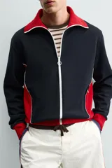 Chaqueta deportiva de corte regular, confeccionada en tejido piqué con interior forrado en red. Presenta un diseño color block en negro y rojo con detalles en blanco, cuello subido, cierre frontal con cremallera y bolsillos laterales con cierre.