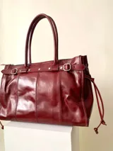 Bolso grande de cuero marrón con textura tipo cocodrilo, estructura rígida y asas para llevar al hombro. Tiene una tira con tachas y hebillas en cada extremo, también lleva lazos en los costados.