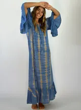Vestido largo de corte holgado con estampado tie-dye en tonos azul y beige. Presenta mangas tres cuartos con volados, escote con cordones y borlas decorativas, y un volado en el ruedo.