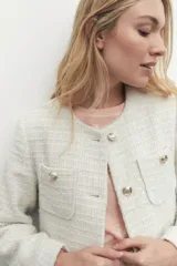 Chaqueta de tweed color crudo con botones metálicos dorados, cuello redondo y bolsillos frontales.