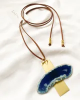 Collar con dije en forma de cruz, compuesto por una placa de latón dorada y una piedra ágata azul irregular. El collar es de cuero marrón con cuentas doradas en los extremos.