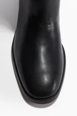 Botas altas de caña recta, color negro, con punta redondeada y taco bajo cuadrado. Presentan una correa decorativa alrededor del tobillo con una hebilla metálica rectangular de color dorado.