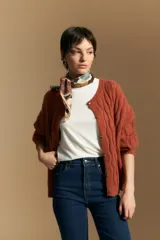 Cardigan de punto color terracota con diseño de trenzas verticales, cuello redondo y cierre frontal con botones metálicos. Presenta mangas largas con puños ajustados y un calce holgado.