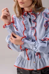 Camisa negra de corte entallado con volados decorativos en el cuello, la botonadura frontal, los hombros y las mangas. Presenta mangas largas con puños ajustados y un ruedo con volado inferior.