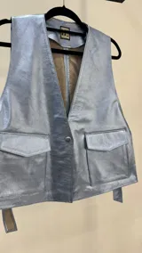 Chaleco corto de efecto metalizado en tono celeste claro, con cuello en V, cierre frontal con un botón a presión y dos bolsillos delanteros con solapa.