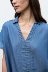 Túnica corta de lyocell color azul jean, con cuello en V, cuello camisero sutil, mangas cortas y costuras con pespuntes visibles en el frente.
