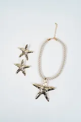 Set compuesto por un collar de cordón trenzado color crema con dije central de estrella de mar dorada y un par de aros a juego con diseño de estrella de mar dorada.