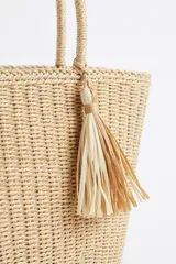 Cartera tipo shopper de mimbre tejido color beige, con doble asa de hombro y detalle de borla colgante.