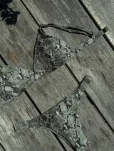 Bikini de dos piezas color marrón, con corpiño triangular fijo con corte bajo el busto, breteles regulables con hebilla y almohadillas desmontables. La bombacha es vedetina con frunce elástico en los costados.