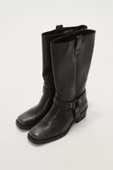 Bota de caña media estilo western, confeccionada en cuero sintético negro. Presenta puntera cuadrada, taco bajo cuadrado y una correa decorativa con tachas metálicas y argolla en el tobillo.