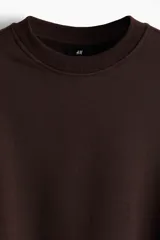 Buzo de cuello redondo color marrón oscuro, confeccionado en tejido de mezcla de algodón con interior cepillado. Presenta un diseño de hombros caídos, corte holgado y terminaciones acanaladas en cuello, puños y bajo.