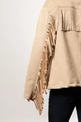 Chaqueta color beige de gamuza sintética con flecos en mangas y canesú, cuello camisero y cierre con botones.