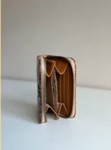 Billetera pequeña de cuero color marrón con textura símil cocodrilo, cierre perimetral en tono rojo y detalles interiores en color tostado. Cuenta con un aplique metálico circular en la parte frontal.