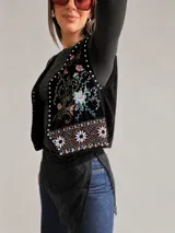 Chaleco de estilo boho chic confeccionado en tela aterciopelada negra, con bordados florales multicolor y detalles de tachas metálicas a lo largo de los bordes frontales.