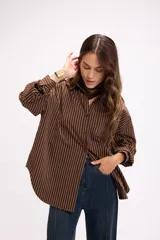 Camisa de corte oversize con estampado de rayas verticales finas en color marrón sobre fondo blanco. Presenta cuello clásico, cierre frontal con botones, bolsillo en el pecho y mangas largas con puños abotonados.