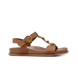 Sandalias planas Korium modelo Corinne, confeccionadas en gamuza marrón, con tiras en forma de T adornadas con tachas y detalles metálicos dorados. Incluyen tira ajustable en el talón con hebilla metálica dorada.