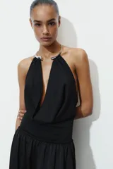 Body negro de cuello halter con detalle de pieza metálica y aplicación joya. Escote pico pronunciado y tejido drapeado. Cierre en bajo con botones a presión.