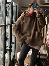 Campera tipo hoodie confeccionada en tela sherpa color verde oliva, con diseño de apliques bordados en la espalda que representan una flor y una fruta. Presenta capucha integrada y corte holgado.