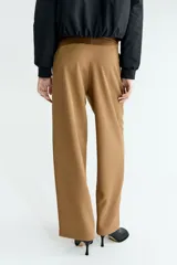Pantalón sastrero de corte holgado en color tostado, con diseño asimétrico cruzado en la cintura, botón frontal y pliegues en las piernas.