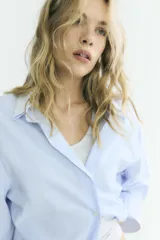 Camisa oversize de algodón con estampado de rayas finas, cuello solapa, escote en pico y manga larga con puños abotonados. Presenta un diseño con pliegues laterales y cierre frontal con botones.