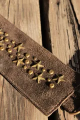Bandolera confeccionada en cuero de gamuza marrón, con cierre superior. Presenta un llavero colgante con forma de estrella revestido en tachas metálicas y tiras flecadas en tono dorado.