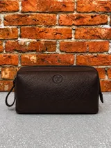 Necessaire de cuero marrón con textura granulada, cierre superior y logo circular grabado en el frente. Incluye una pequeña manija lateral para su transporte.