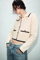 Chaqueta de punto color crema con ribetes en contraste negro, cuello solapa, manga larga y cierre frontal con botones dorados. Presenta bolsillos de plastrón en el delantero con detalle de botones.