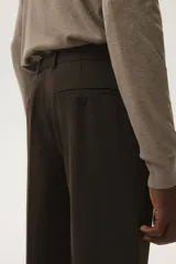 Pantalón de vestir color marrón oscuro, corte slim fit, con pliegue marcado en la pierna y puños doblados. Presenta cierre frontal con botón y bolsillos laterales y traseros.