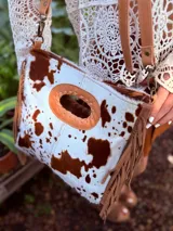 Bandolera de cuero con estampado animal print de vaca en tonos marrón y blanco, con flecos de cuero marrón en los laterales. Tiene correa larga de cuero marrón regulable y asa de mano con forma ovalada.