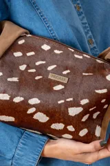 Cartera de hombro con diseño de animal print tipo vaca en tonos marrón y blanco. Presenta una correa ancha de gamuza marrón y una placa metálica con logo en el frente.